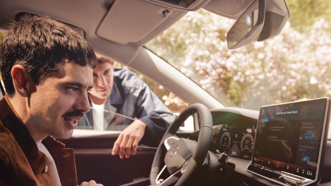 Ein Mann sitzt im neuen VW Golf und schaut auf das Infotainment-Display, ein anderer schaut durch das Fenster.