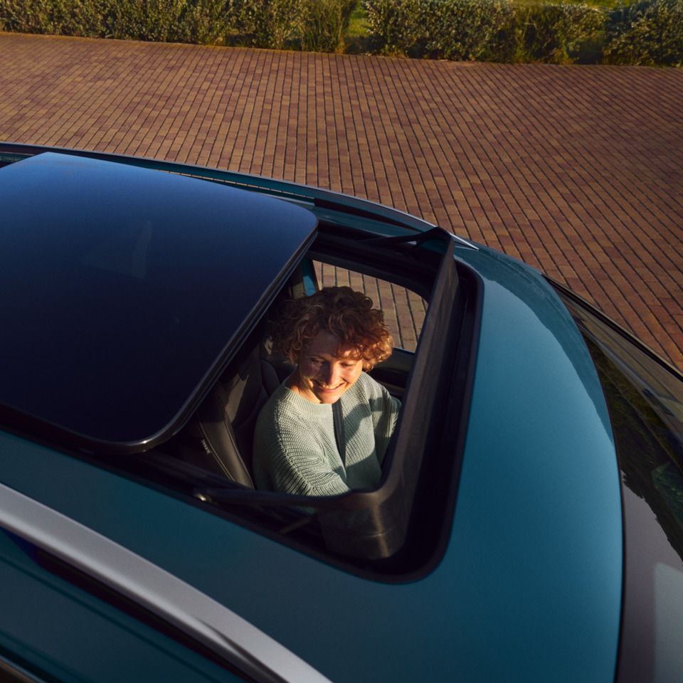  Blick von oben auf blauen VW Tiguan mit offenem Panorama-Ausstell-/Schiebedach, eine Frau schaut von innen heraus.