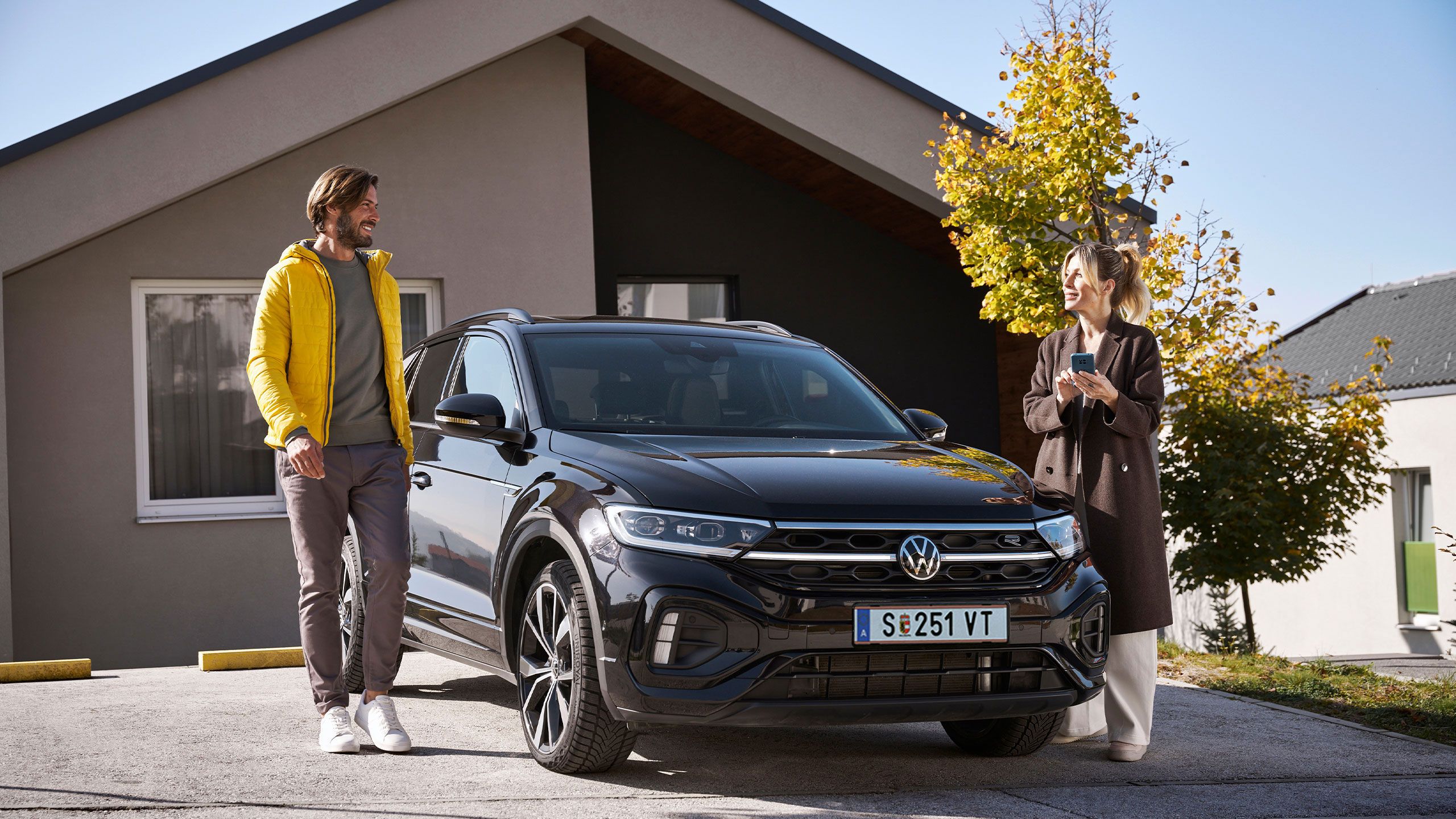 Mann und Frau steht mit Smartphone neben einem geparkten T-Roc vor einem Haus.