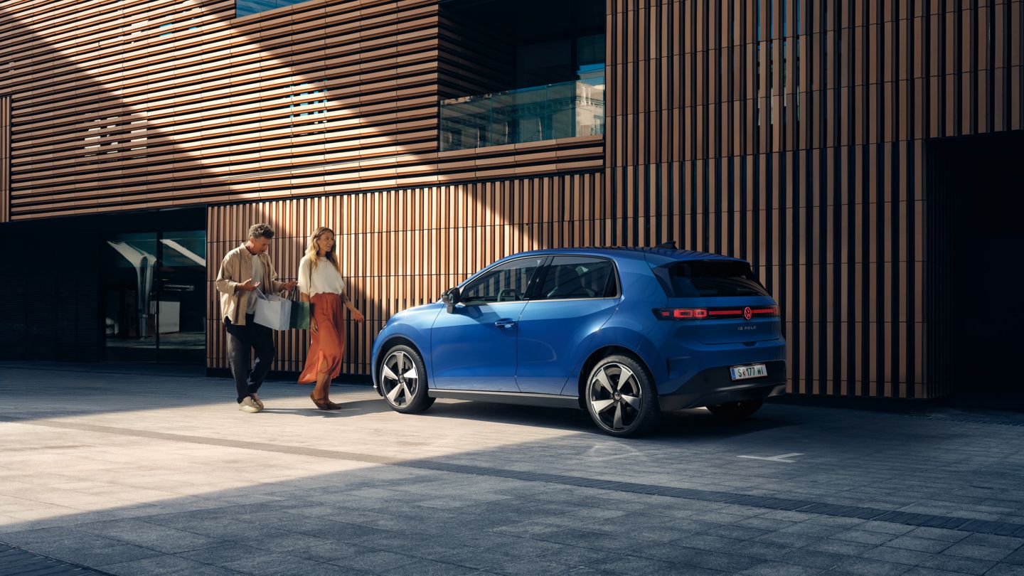 Ein blauer VW ID. Polo Style steht vor einem Gebäude mit Holzfassade. Ein Mann und eine Frau mit Einkaufstaschen gehen darauf zu