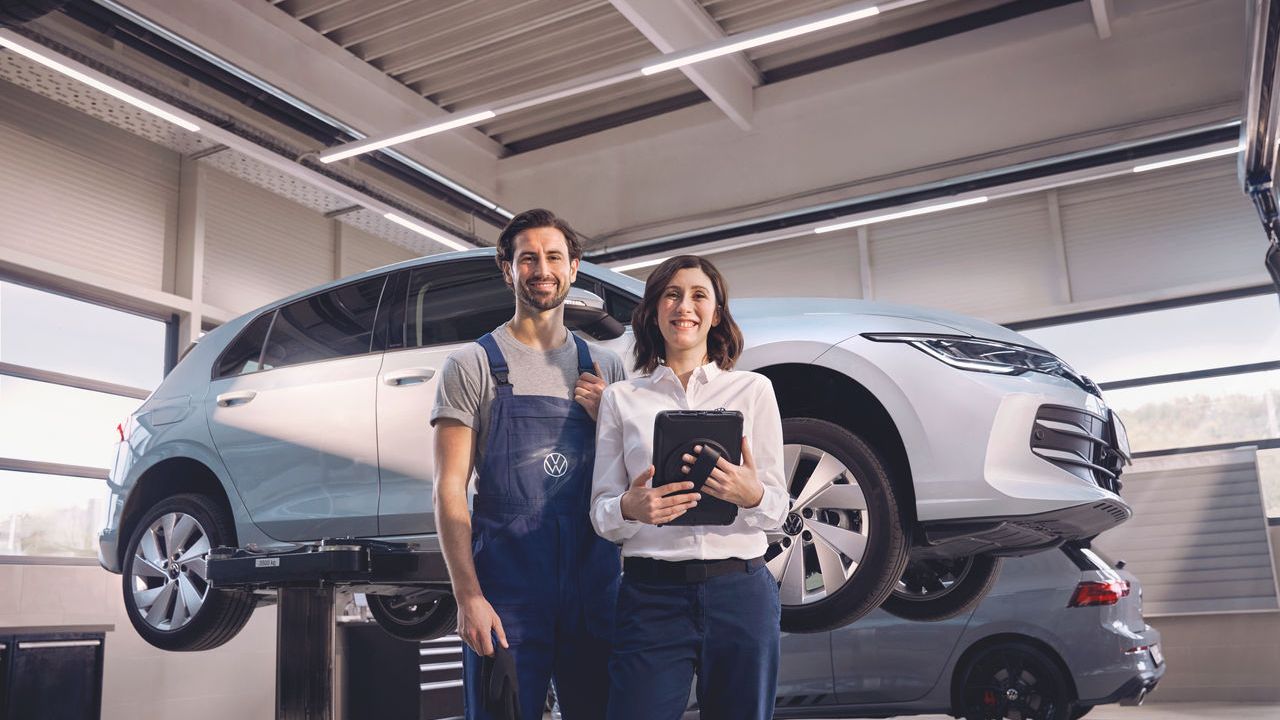 Zwei Volkswagen Mitarbeiter:innen lachen in die Kamera. Im Hintergrund ein Volkswagen Golf auf einer Hebebühne.