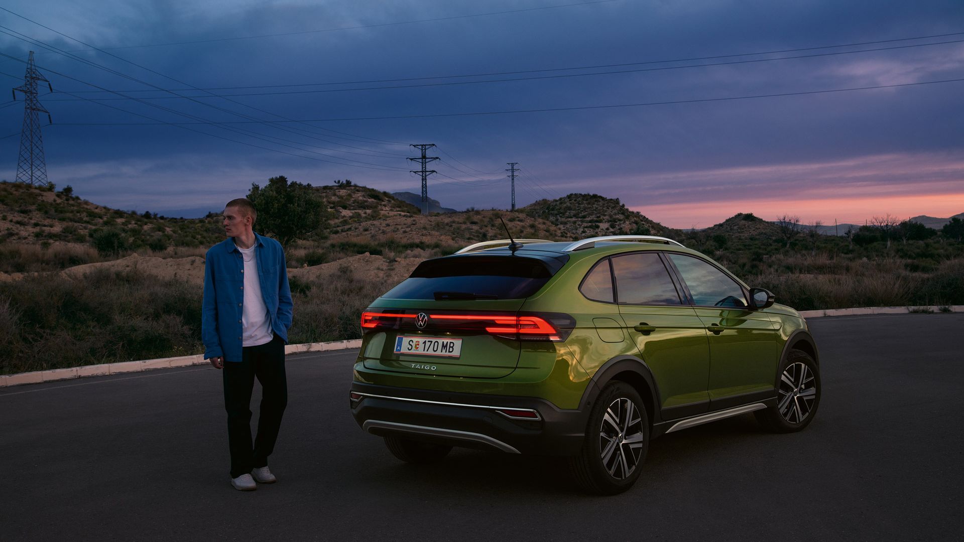 Ein Mann steht hinter einem grünen VW Taigo mit eingeschaltetem Licht auf einem Wüstenparkplatz im Sonnenuntergang.