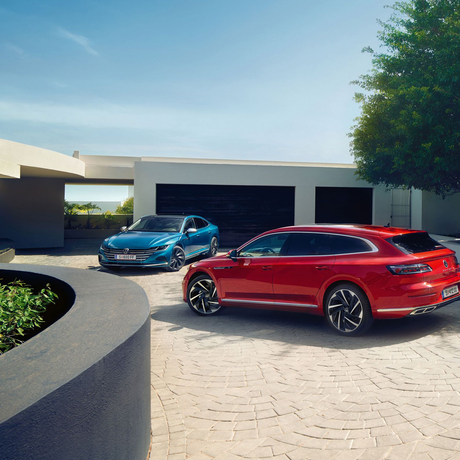 Zwei Arteon sind vor einem eleganten Haus mit weißer Fassade und schwarzen Garagentoren geparkt. Ein rotes Auto steht im Vordergrund, während ein blaues Auto im Hintergrund nahe der Garage parkt. Die Szene ist von klarem Himmel und grünen Pflanzen umgeben.