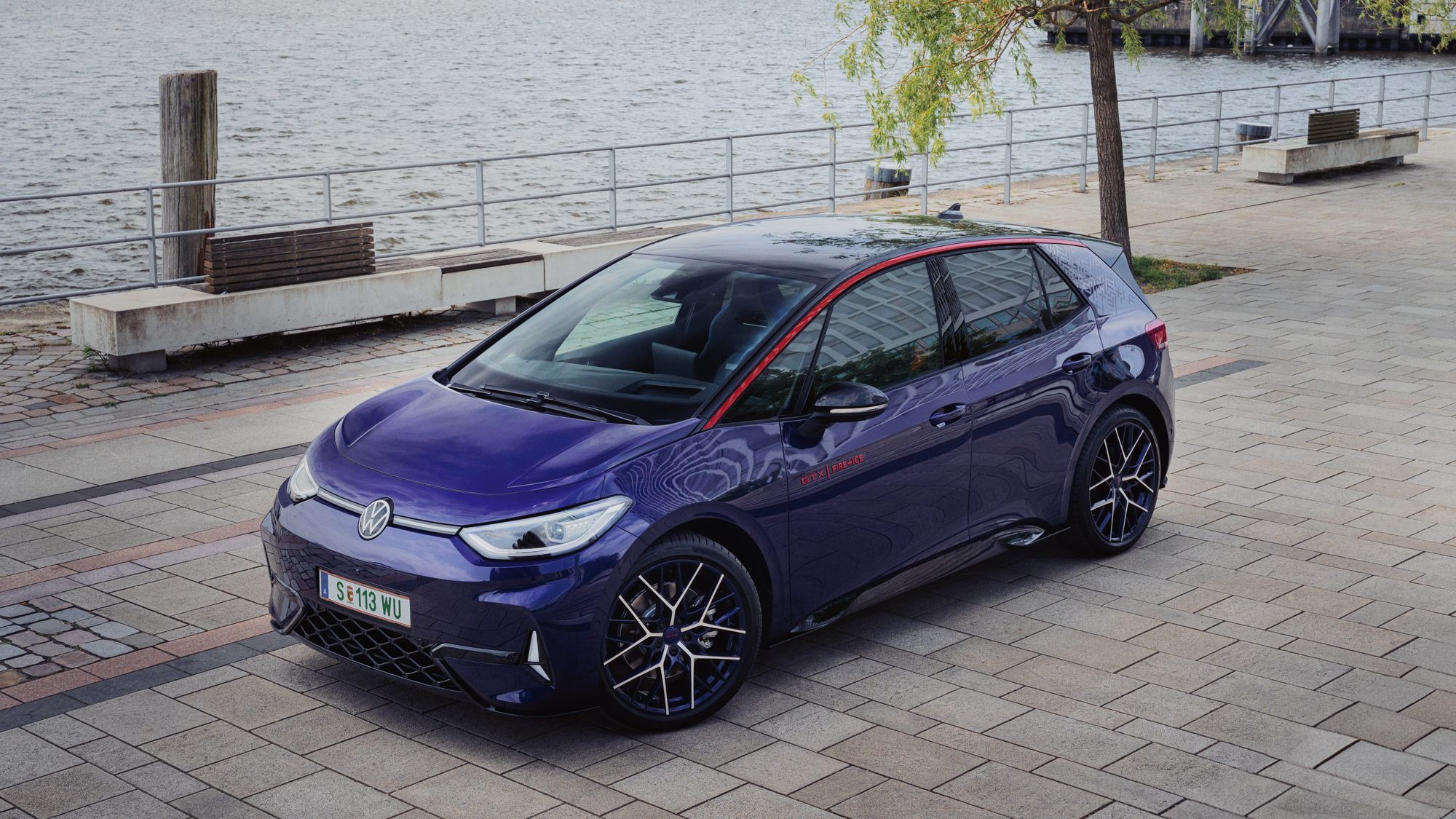 Ein blauer Volkswagen ID.3 GTX steht auf einem gepflasterten Platz am Ufer eines Gewässers. Der Hintergrund zeigt Bäume und industrielle Strukturen. Das Auto hat schwarze Felgen und rote Akzente.
