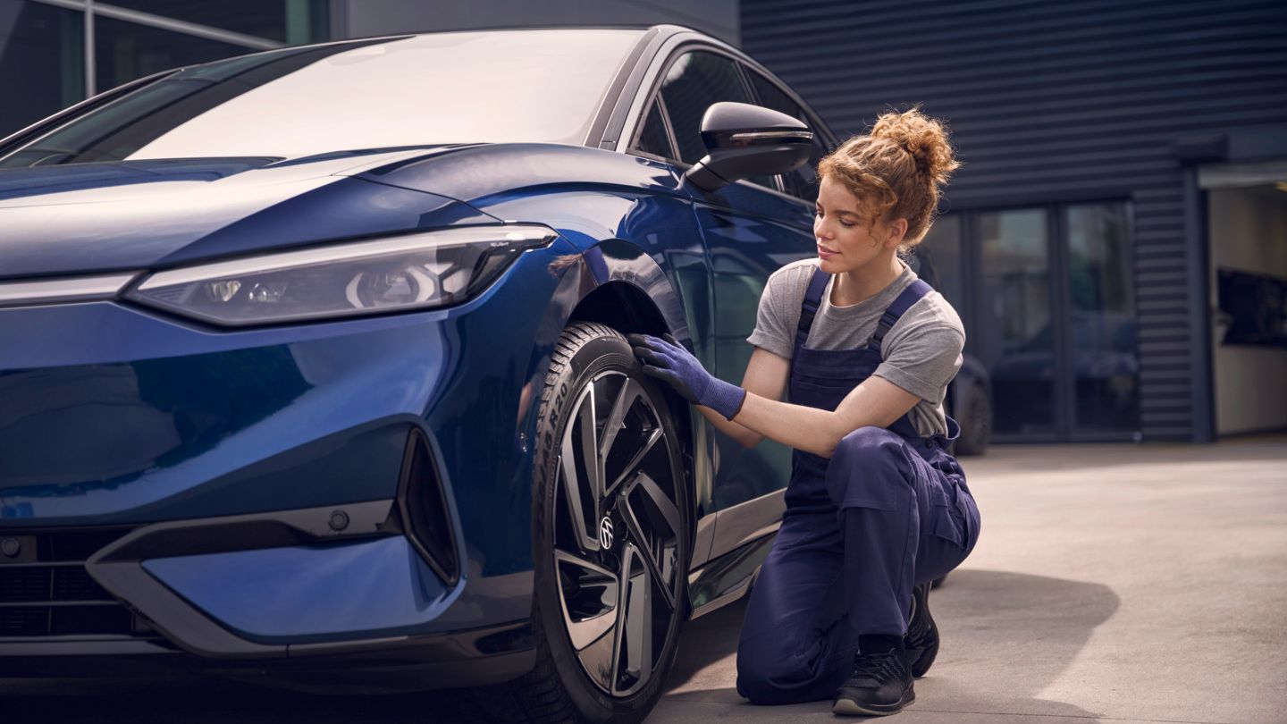 Eine Frau im blauen Overall kniet neben einem blauen VW ID.7 und prüft den linken Vorderreifen.