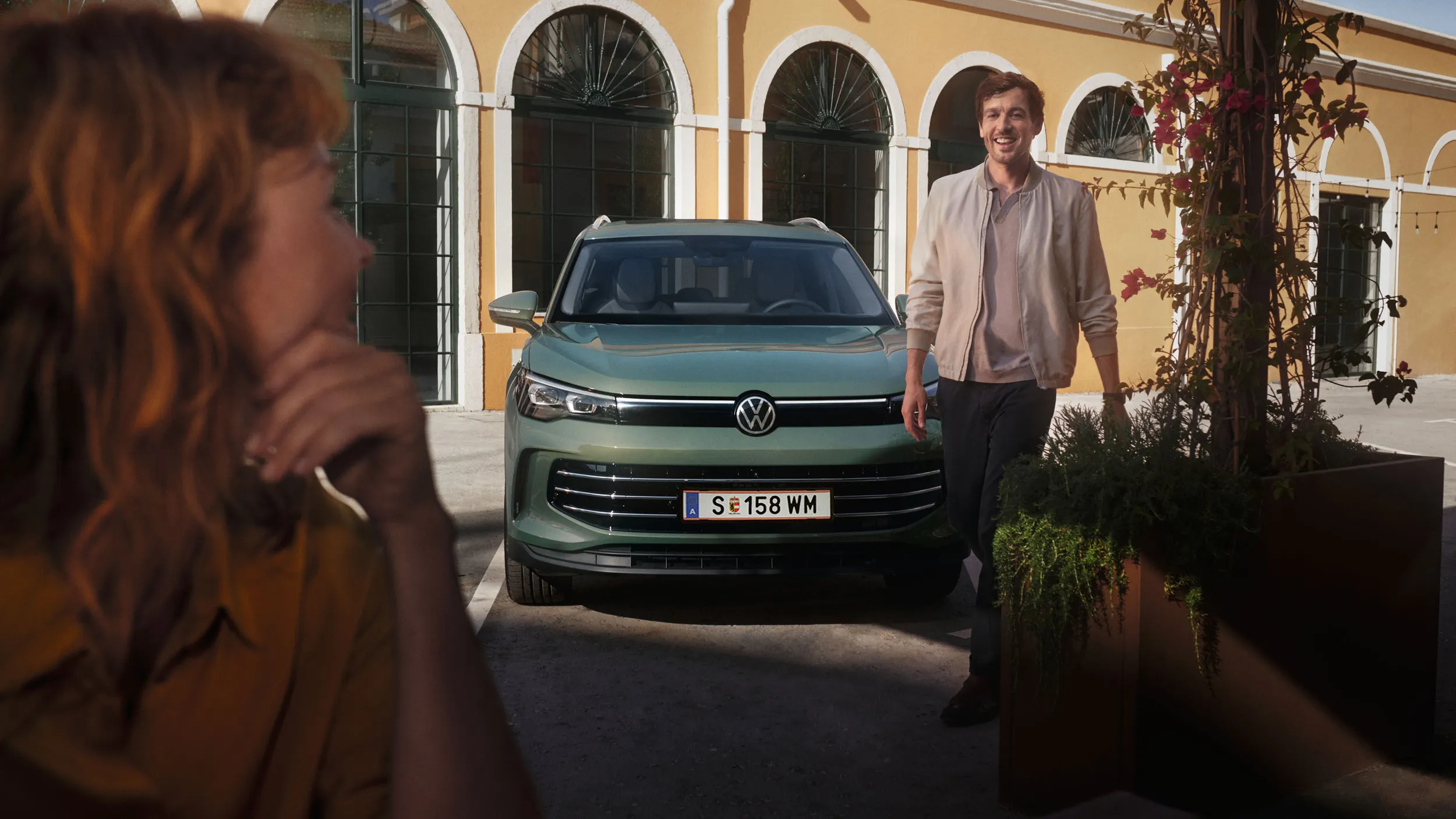 Tiguan ist vor einem Haus auf einem Parkplatz geparkt von dem ein Mann weggeht und ihn im Vordergrund währenddessen eine Frau anssieht.