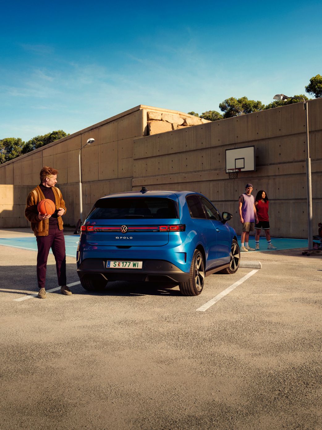 Ein blauer VW ID. Polo parkt vor einem Basketballplatz, daneben steht ein Mann mit Basketball in der Hand.
