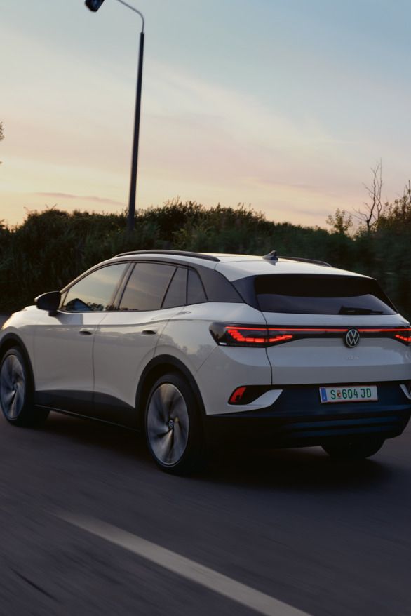 Ein weißer VW ID.4 fährt auf einer Landstraße bei Sonnenuntergang. Das Auto ist von hinten zu sehen. Die Straße ist von Bäumen und Sträuchern gesäumt.