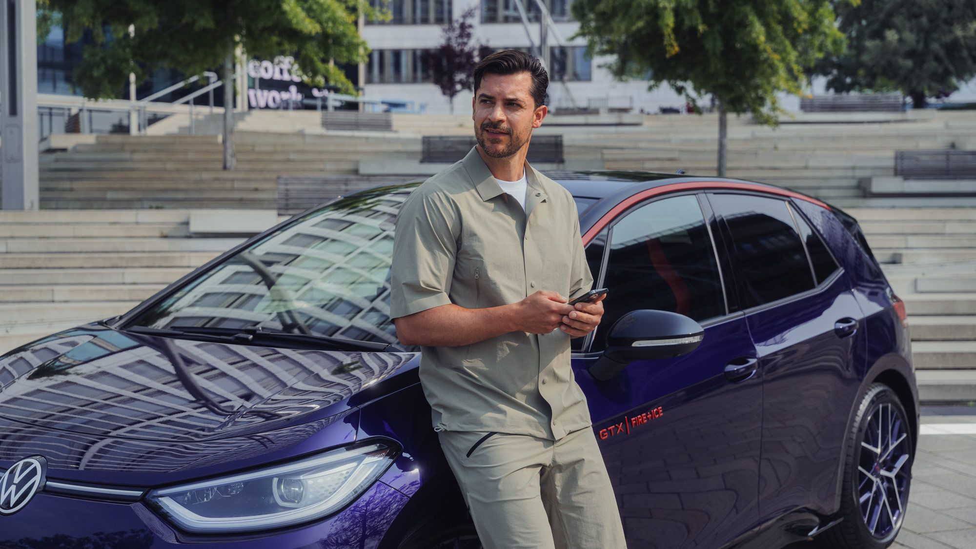 Ein Mann lehnt lässig an einem lilafarbenem Volkswagen ID.3 GTX FIRE+ICE. Der Mann trägt ein beigefarbenes Hemd und Shorts und hält ein Smartphone in der Hand. Im Hintergrund sind moderne Gebäude und Treppenstufen zu sehen.