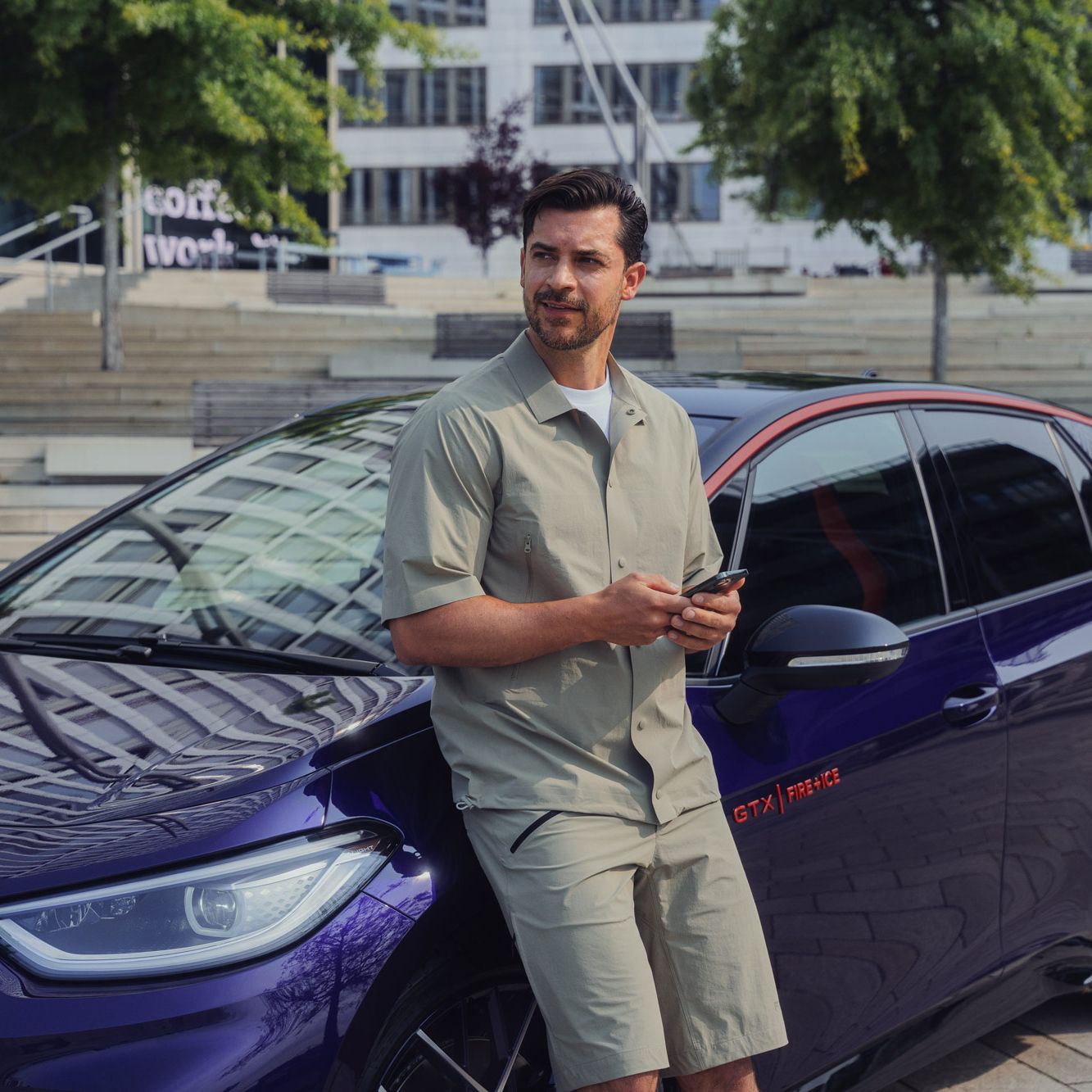 Ein Mann lehnt lässig an einem lilafarbenem Volkswagen ID.3 GTX FIRE+ICE. Der Mann trägt ein beigefarbenes Hemd und Shorts und hält ein Smartphone in der Hand. Im Hintergrund sind moderne Gebäude und Treppenstufen zu sehen.