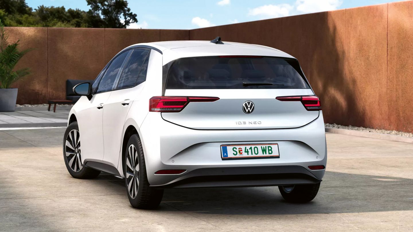 Ein weißer Volkswagen ID.3 Neo steht auf einem modernen, sonnigen Innenhof vor einer niedrigen, rostfarbenen Mauer. Das Auto ist von hinten leicht schräg zu sehen, mit klar erkennbaren LED-Rückleuchten und österreichischem Kennzeichen. Im Hintergrund sind ein paar Pflanzen und ein blauer Himmel mit leichten Wolken zu sehen.