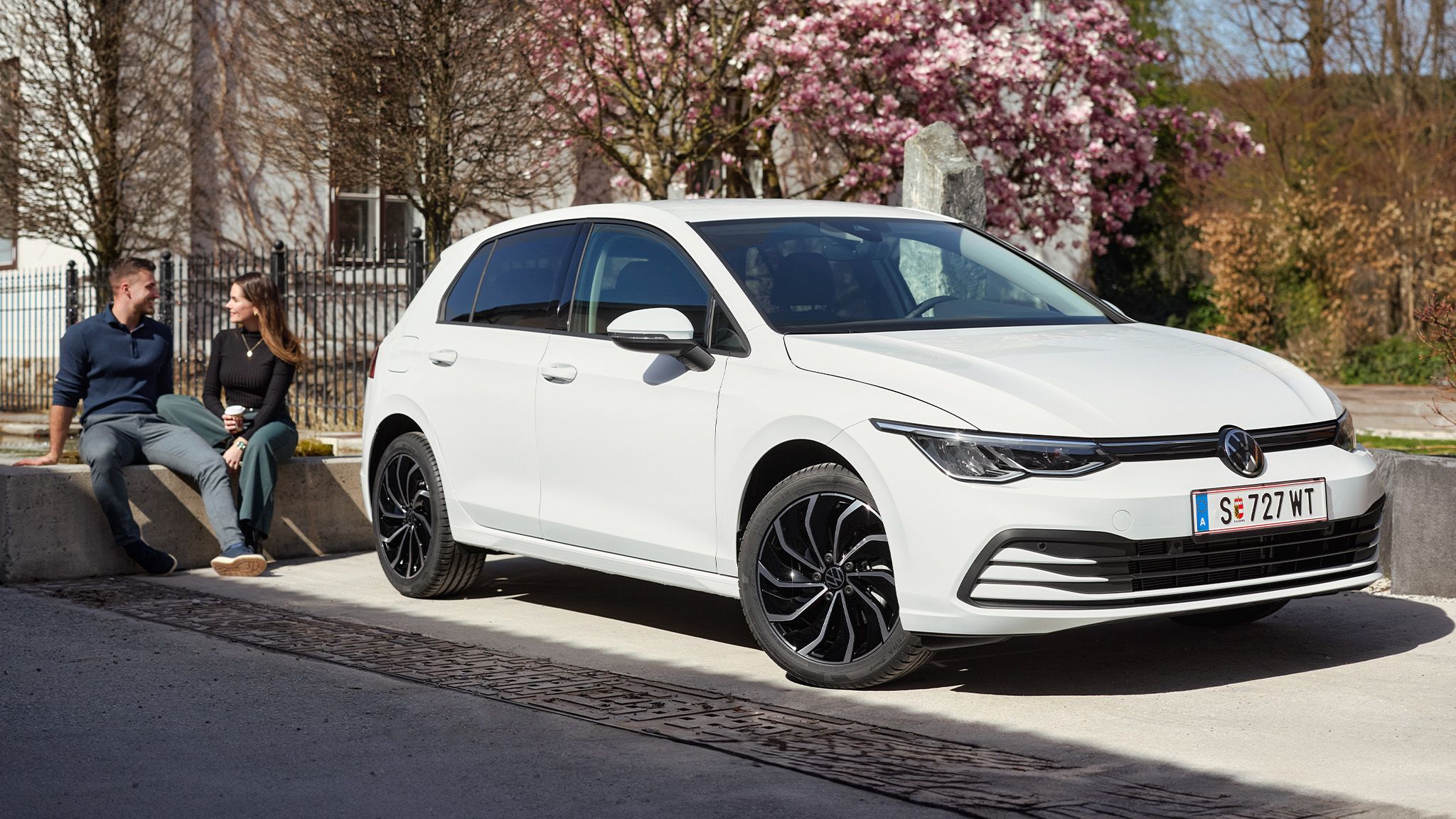 Ein weißer Volkswagen Golf steht vor einem blühenden Baum. Im Hintergrund sind zwei Personen, die auf einer Betonkante sitzen, leicht verschwommen zu sehen. Die Szene befindet sich im Freien an einem sonnigen Tag.