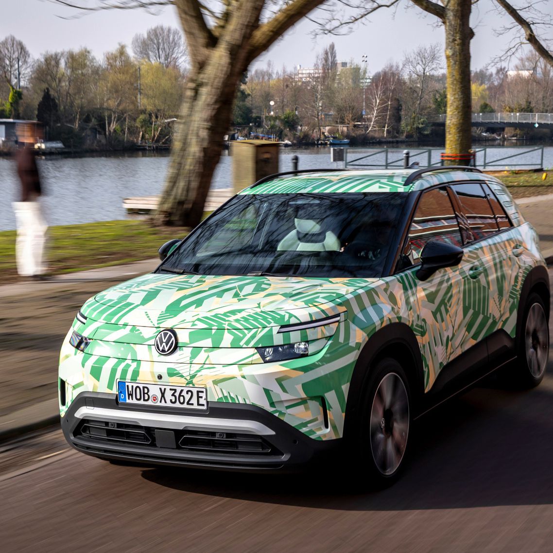 Ein Elektroauto fährt auf einer Straße entlang eines Flusses. Es hat eine grüne Tarnlackierung und moderne LED-Scheinwerfer. Im Hintergrund sind Bäume und eine Brücke zu sehen.