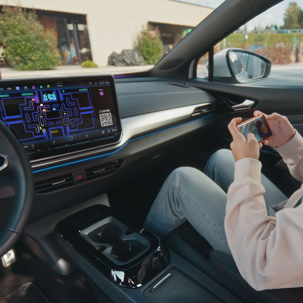 Eine Person sitzt entspannt im Fahrersitz eines Autos und spielt ein Videospiel auf ihrem Smartphone. Das Auto hat ein großes eingebautes Display auf dem Armaturenbrett, das ebenfalls ein Videospiel zeigt. Im Hintergrund ist die Außenseite eines Gebäudes zu sehen.
