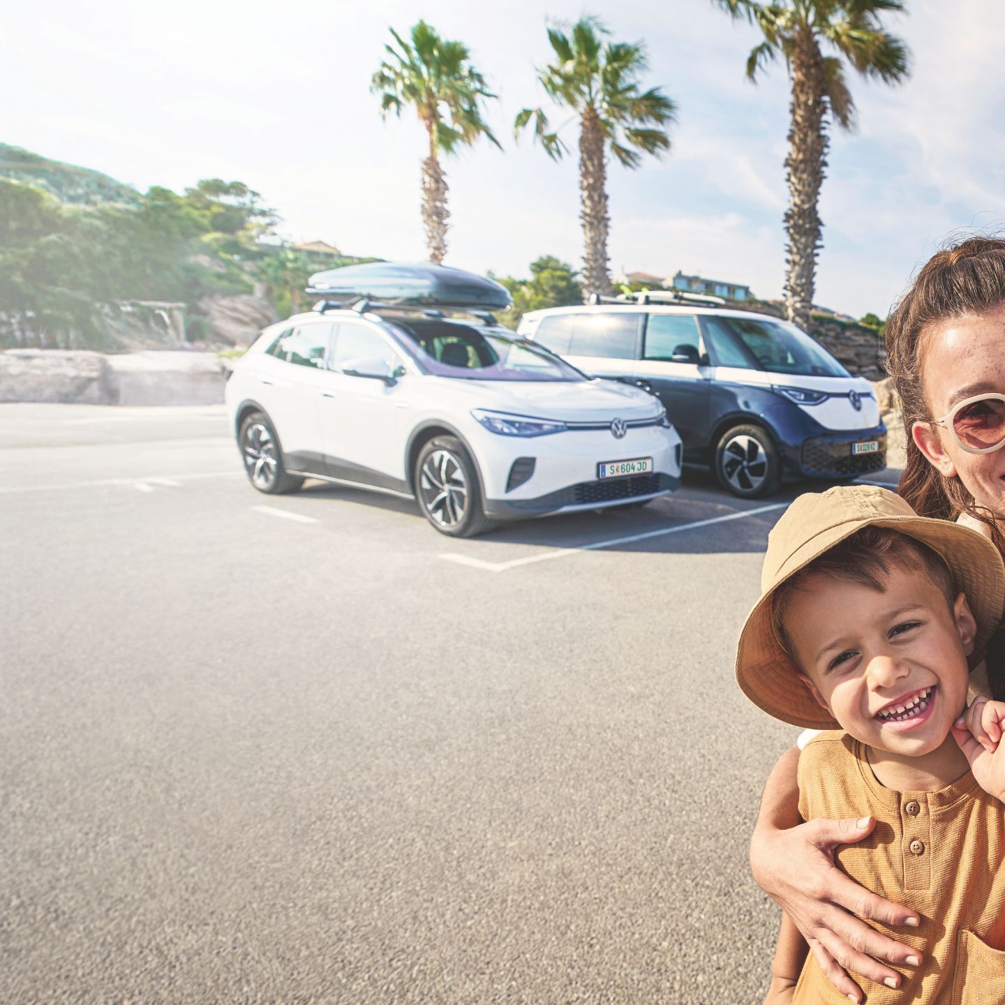 Eine Familie macht ein Selfie auf einem sonnigen Parkplatz mit Palmen im Hintergrund. Im Hintergrund stehen zwei geparkte Volkswagen mit Gepäck auf dem Dach.