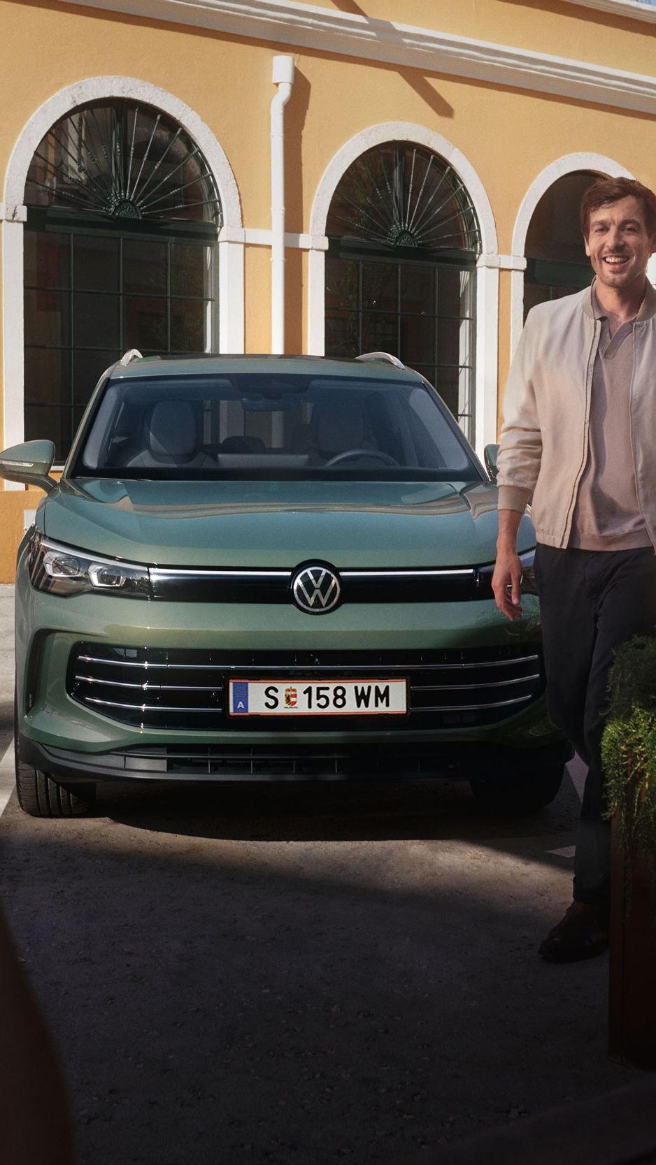 Tiguan ist vor einem Haus auf einem Parkplatz geparkt von dem ein Mann weggeht und ihn im Vordergrund währenddessen eine Frau anssieht.
