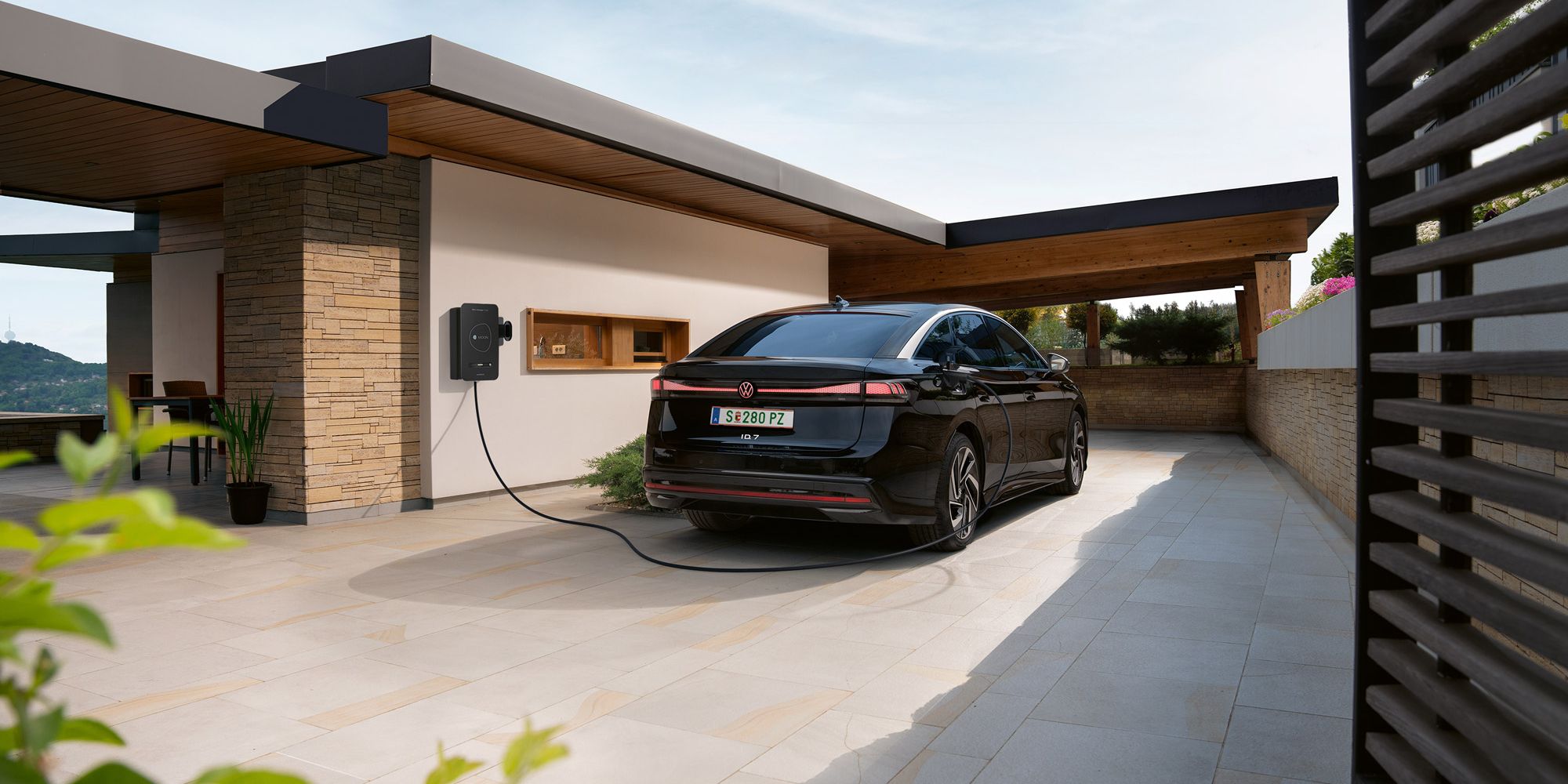 Ein schwarzes Elektroauto steht in einer modernen Einfahrt und ist an einer Wallbox an der Hauswand zum Laden angeschlossen. Das flache, kubische Haus mit Holz- und Steinelementen wirkt modern und hochwertig. Im Vordergrund sind Pflanzen zu sehen, im Hintergrund öffnet sich der Blick auf eine Landschaft.