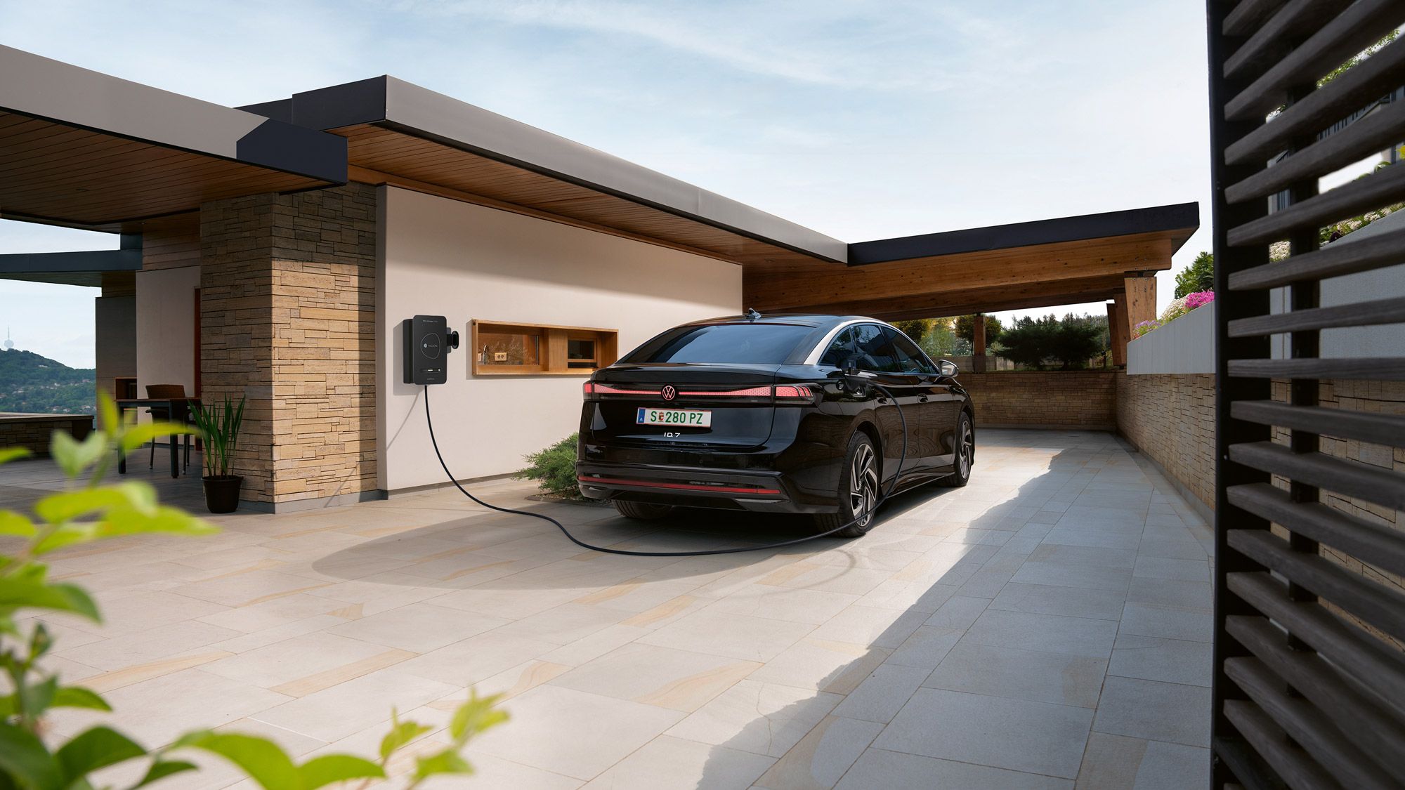 Ein schwarzes Elektroauto steht in einer modernen Einfahrt und ist an einer Wallbox an der Hauswand zum Laden angeschlossen. Das flache, kubische Haus mit Holz- und Steinelementen wirkt modern und hochwertig. Im Vordergrund sind Pflanzen zu sehen, im Hintergrund öffnet sich der Blick auf eine Landschaft.