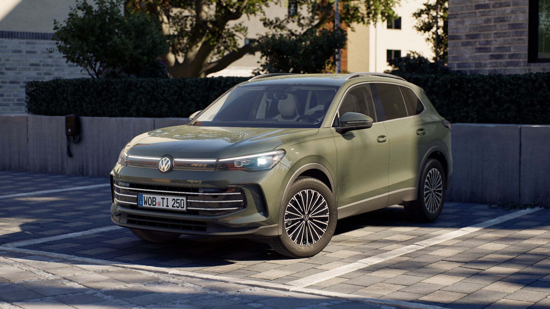 Ein olivgrüner Tiguan von Volkswagen steht auf einem gepflasterten Parkplatz vor modernen Wohngebäuden. Das Auto ist frontal leicht seitlich zu sehen, mit LED-Scheinwerfern und markanten, filigranen Felgen. Im Hintergrund stehen Bäume und eine niedrige Mauer, die eine ruhige Wohngegend andeuten.