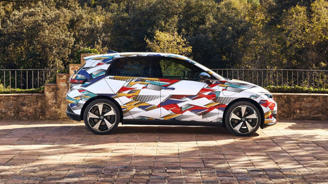 Ein ID. Polo mit buntem, geometrisch gemustertem Aufdruck steht auf einem gepflasterten Platz. Hinter dem Auto erstreckt sich eine bewaldete, hügelige Landschaft. Die Sonne scheint, und das Wetter ist klar.