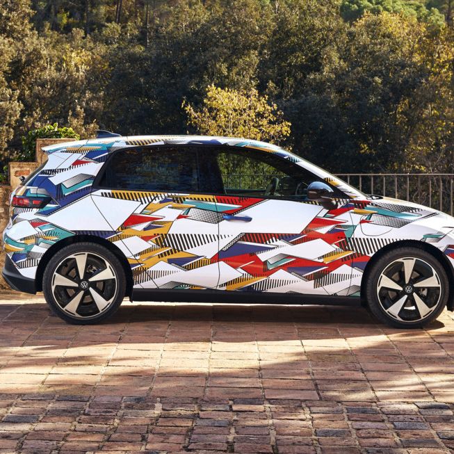 Ein ID. Polo mit buntem, geometrisch gemustertem Aufdruck steht auf einem gepflasterten Platz. Hinter dem Auto erstreckt sich eine bewaldete, hügelige Landschaft. Die Sonne scheint, und das Wetter ist klar.