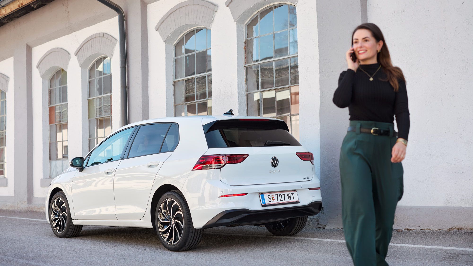 Eine Frau, geht telefonierend neben einem weißen VW Golf, der vor einem Gebäude mit großen Fenstern geparkt ist.