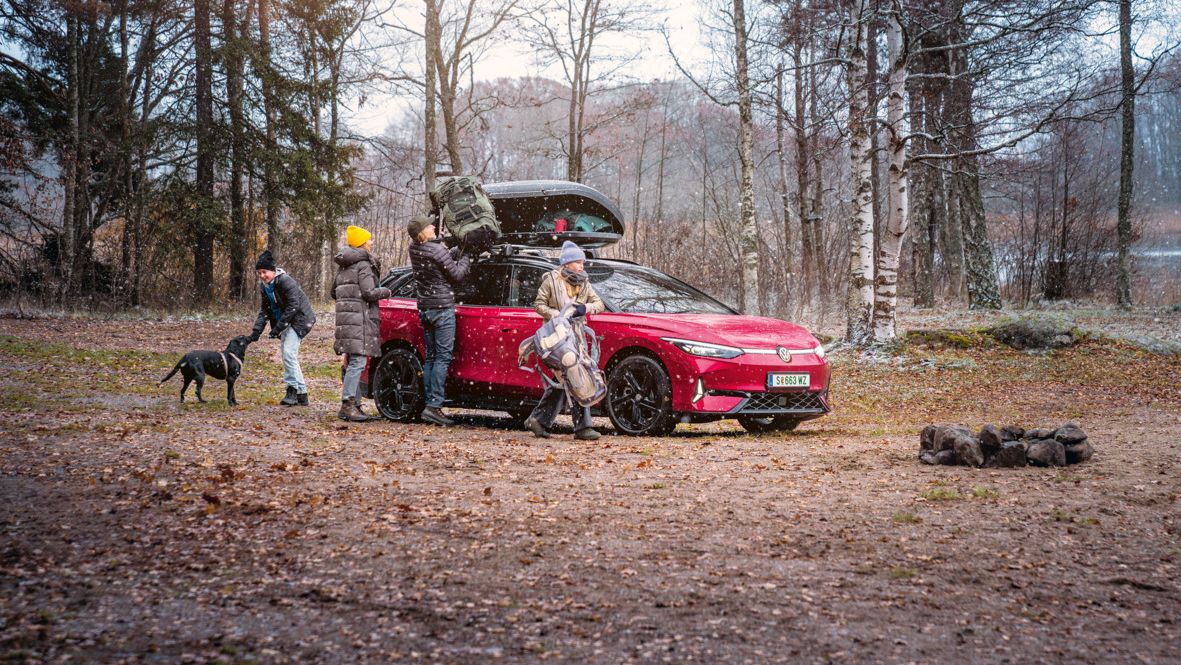 Eine Familie in Winterkleidung entlädt die Dachbox eines roten VW ID.7 GTX Tourer, der am Wald parkt.