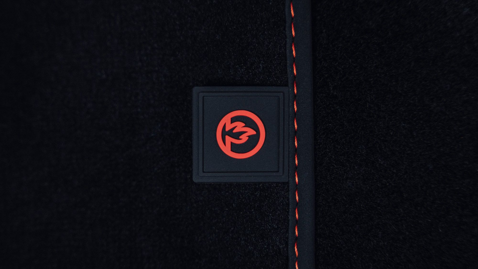 Ein schwarzes Stoffmaterial mit einer roten Naht auf der rechten Seite. Im Zentrum des Bildes befindet sich ein quadratisches Logo mit dem roten Symbol eines stilisierten Feuers. Der Hintergrund ist einfarbig schwarz.