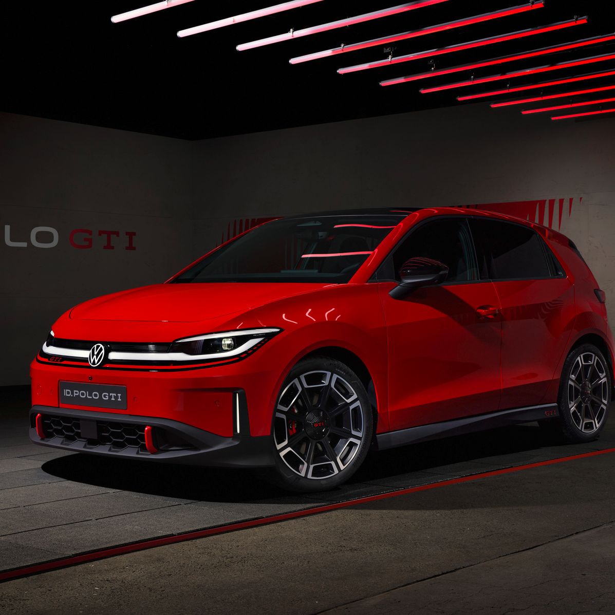 Roter Volkswagen ID. Polo GTI steht in einer dunklen, modernen Garage mit roten Lichtstreifen an der Decke. Das Auto wirkt sportlich mit tiefem Frontspoiler, markanten LED-Scheinwerfern und großen, grauen Leichtmetallfelgen. An der Wand im Hintergrund ist der Schriftzug „ID. POLO GTI“ zu sehen.