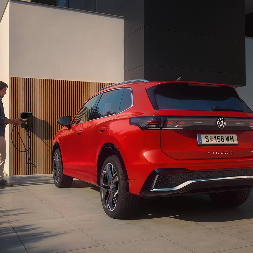Roter VW Tiguan eHybrid steht vor einem modernen Wohnhaus. Ein Mann daneben hält den Stecker einer Ladestation in der Hand.