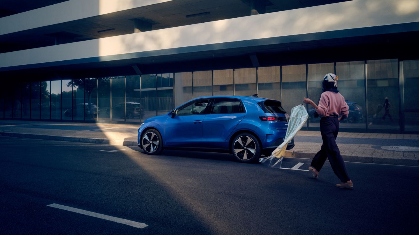 Ein blauer VW ID. Polo Style steht seitlich parkend am Straßenrand, eine Frau geht auf das Fahrzeug zu.