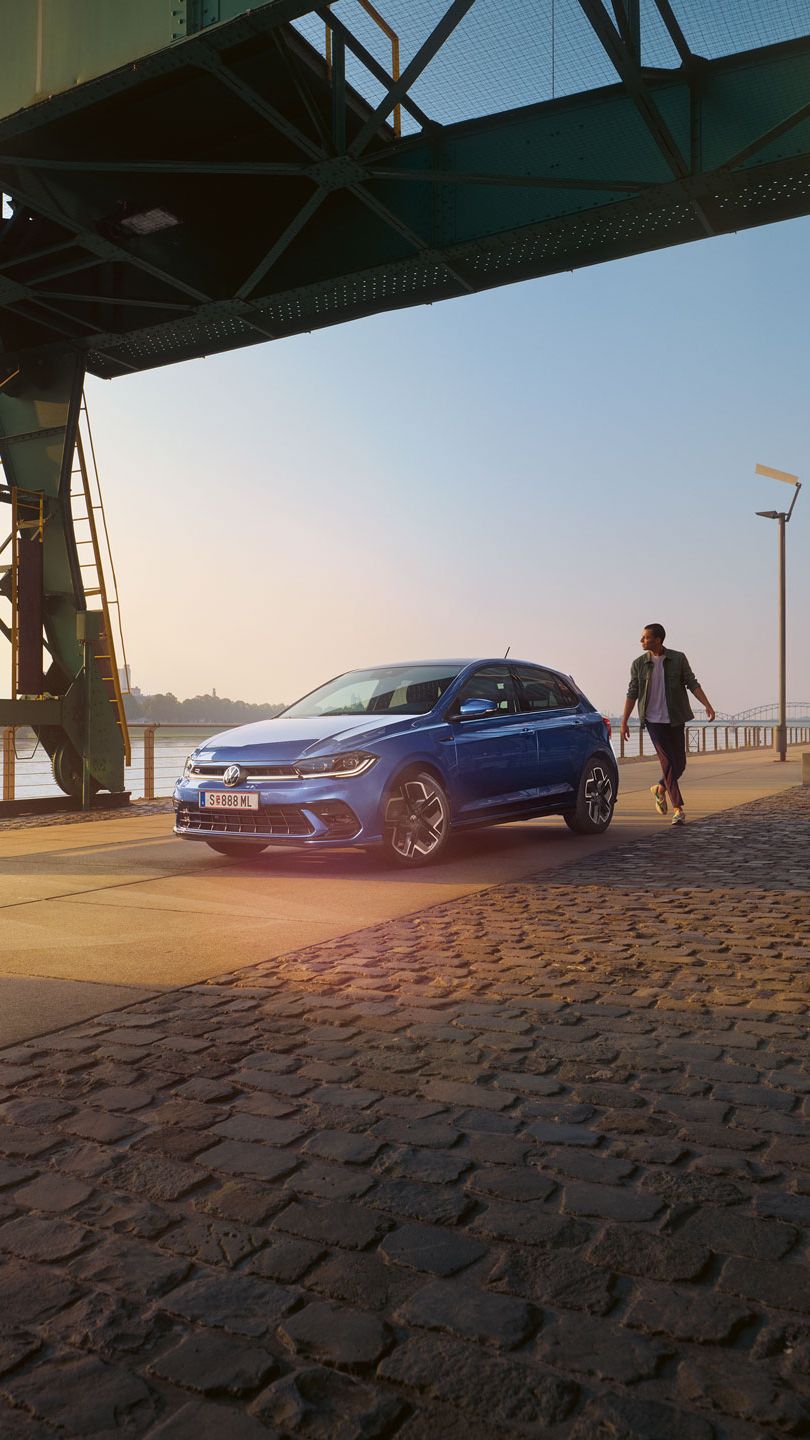  Ein blauer VW Polo R-Line steht an der Promenade neben einem Fluss, ein Mann läuft um das Auto herum.
