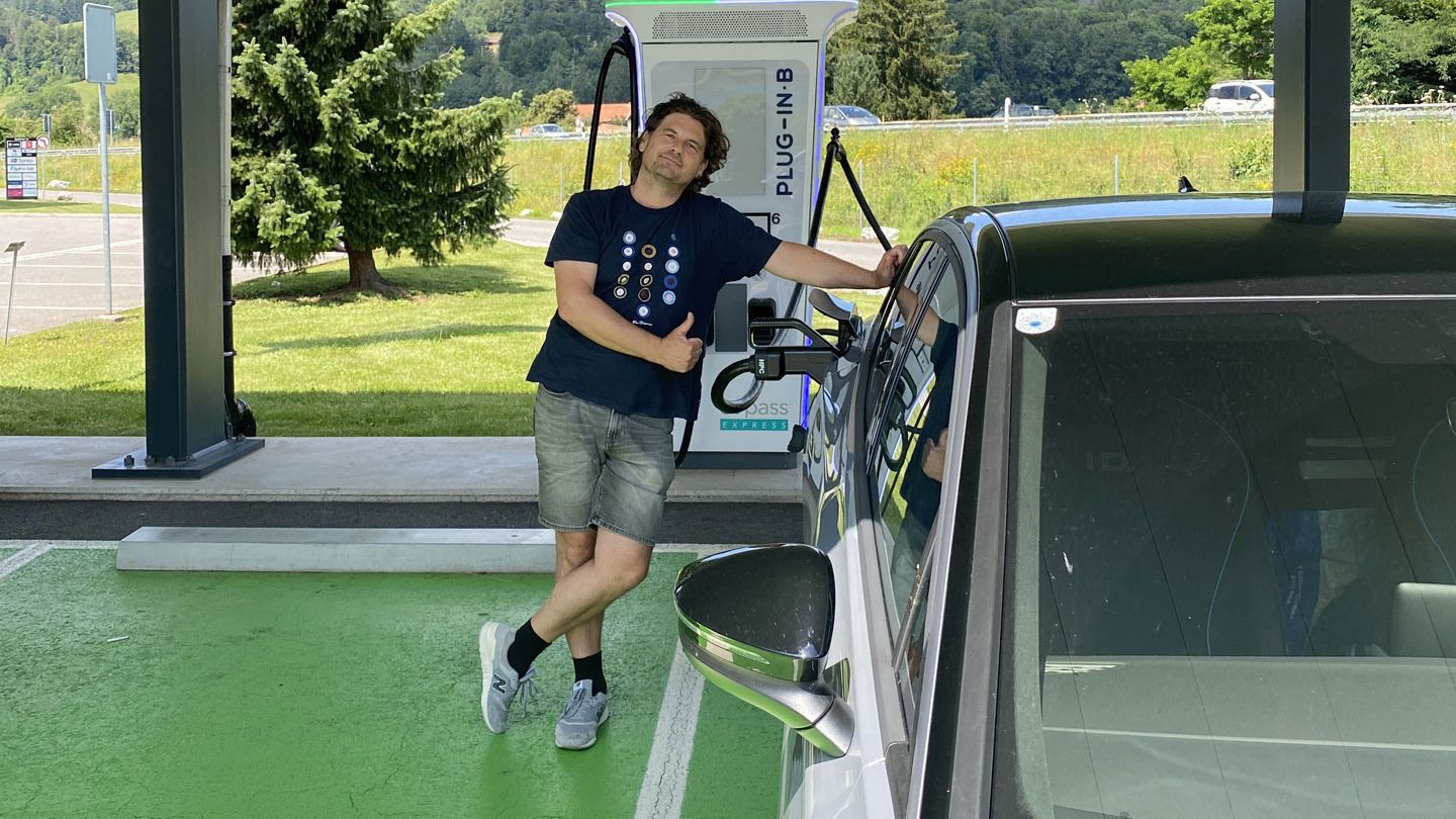 Eine Person lehnt sich an ein Elektroauto, das an einer Ladestation angeschlossen ist. Sie zeigt einen Daumen hoch. Im Hintergrund befinden sich Bäume und Wiesen.