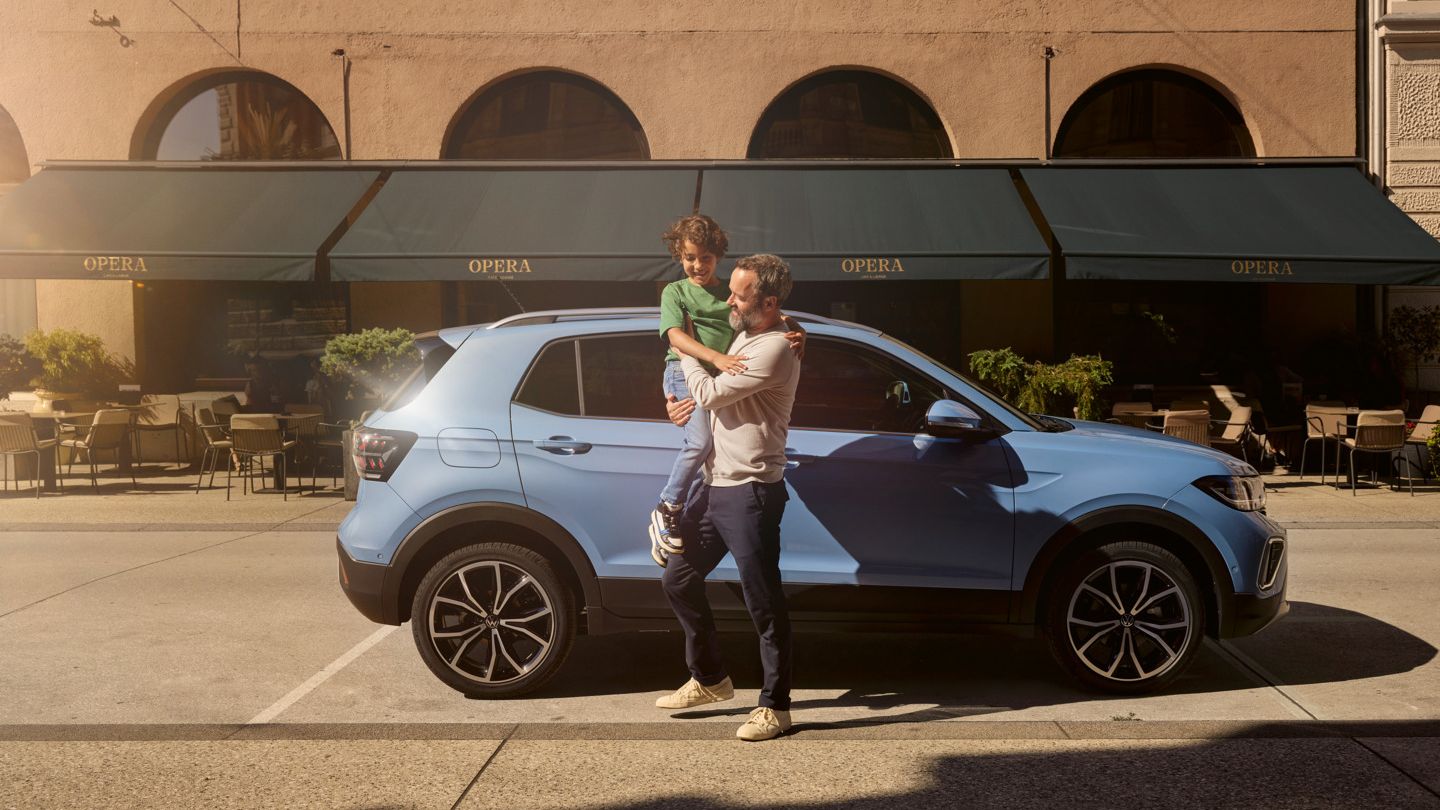 Ein Mann hält einen fröhlichen Jungen vor einem blauen VW T-Cross auf dem Arm.