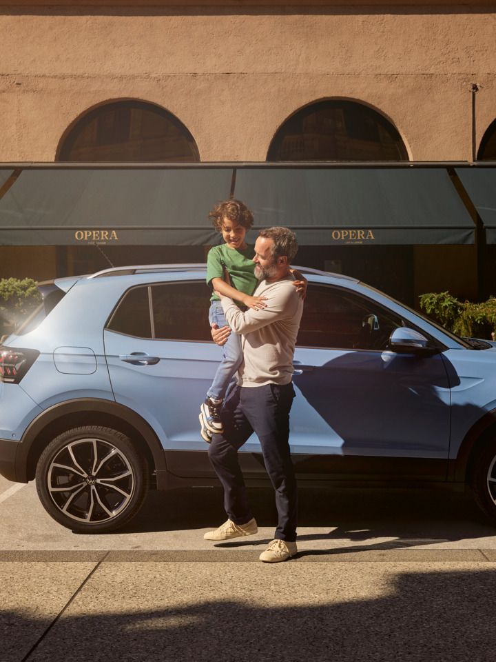 Ein Mann hält einen fröhlichen Jungen vor einem blauen VW T-Cross auf dem Arm.