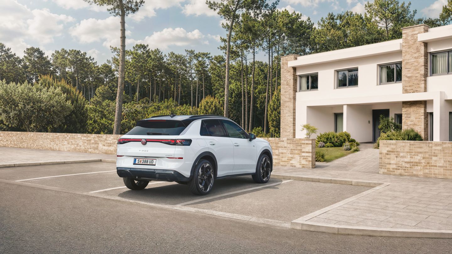 Ein weißer VW T-Roc Hybrid steht auf einem Parkplatz vor einem Wohnhaus am Wald, Blick auf das Heck.