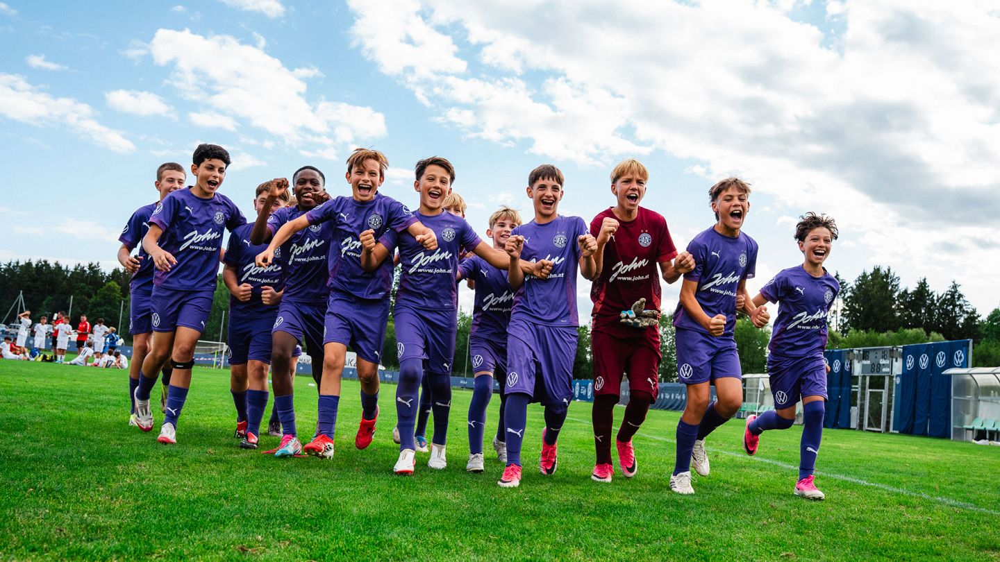 Eine Gruppe jubelnder Kinder in lila Fußballtrikots läuft lachend über einen grünen Rasenplatz. Sie halten sich an den Schultern oder Händen und wirken ausgelassen und fröhlich. Im Hintergrund sind weitere Spieler, Gebäude und ein teilweise bewölkter Himmel zu sehen.