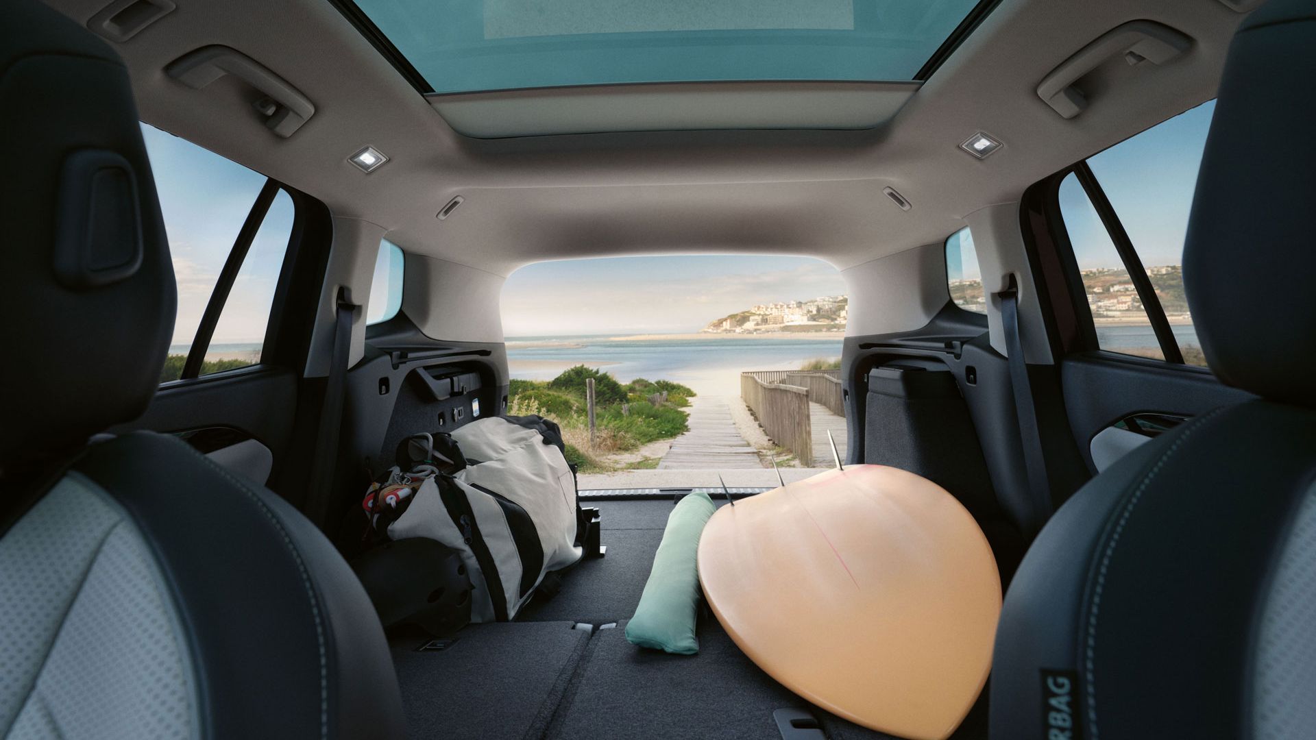 Surfbrett im Kofferraum eines VW Tayron mit umgeklappten Rücksitzen, durch die offene Heckklappe Strand und Meer sichtbar.