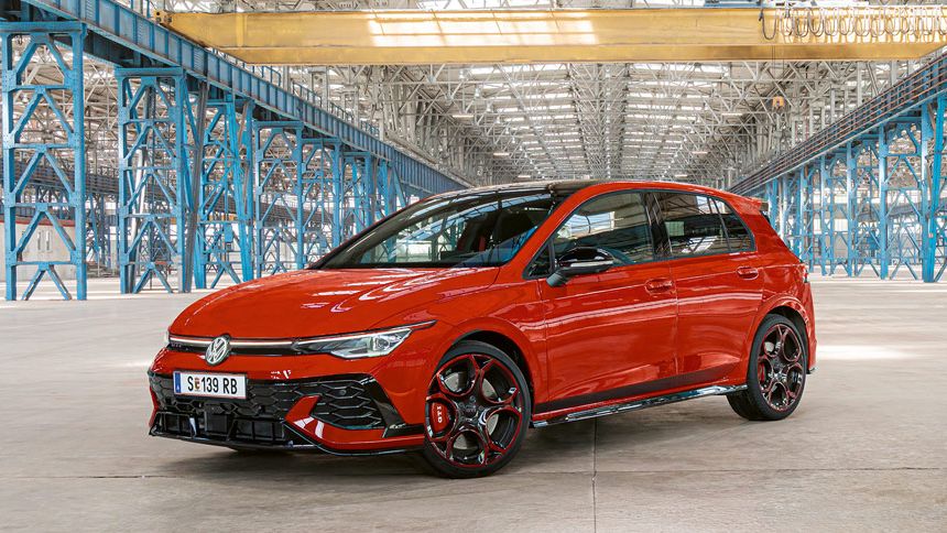 Ein roter Golf GTI steht in einer großen, leeren Industriehalle mit blauen Stahlträgern. Das Dach der Halle ist aus Glas und lässt viel Licht herein.