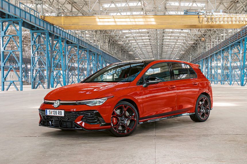 Ein roter Golf GTI steht in einer großen, leeren Industriehalle mit blauen Stahlträgern. Das Dach der Halle ist aus Glas und lässt viel Licht herein.