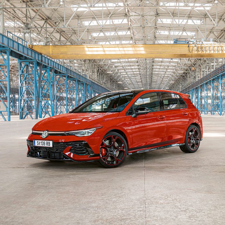 Ein roter Golf GTI steht in einer großen, leeren Industriehalle mit blauen Stahlträgern. Das Dach der Halle ist aus Glas und lässt viel Licht herein.