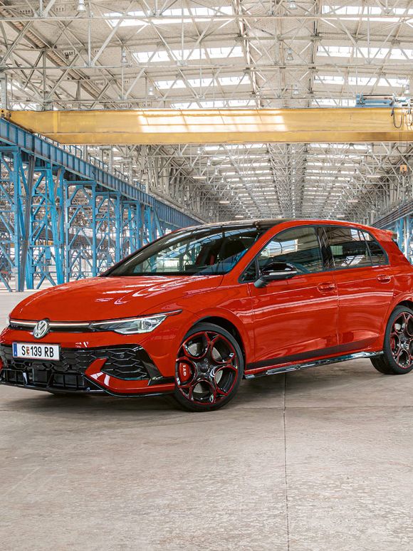 Ein roter Golf GTI steht in einer großen, leeren Industriehalle mit blauen Stahlträgern. Das Dach der Halle ist aus Glas und lässt viel Licht herein.