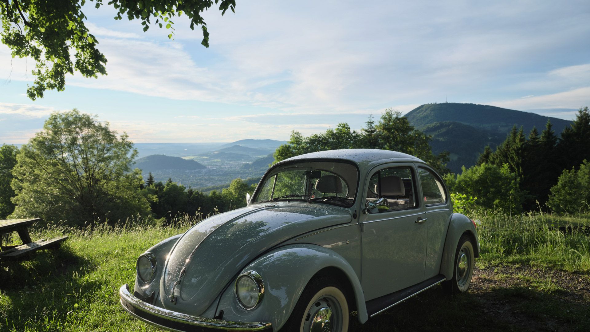Ein grauer VW Käfer steht auf einer grünen Wiese. Im Hintergrund sind Bäume und hügelige Landschaften unter einem bewölkten Himmel zu sehen. Die Sonne scheint durch die Wolken und beleuchtet das Auto.
