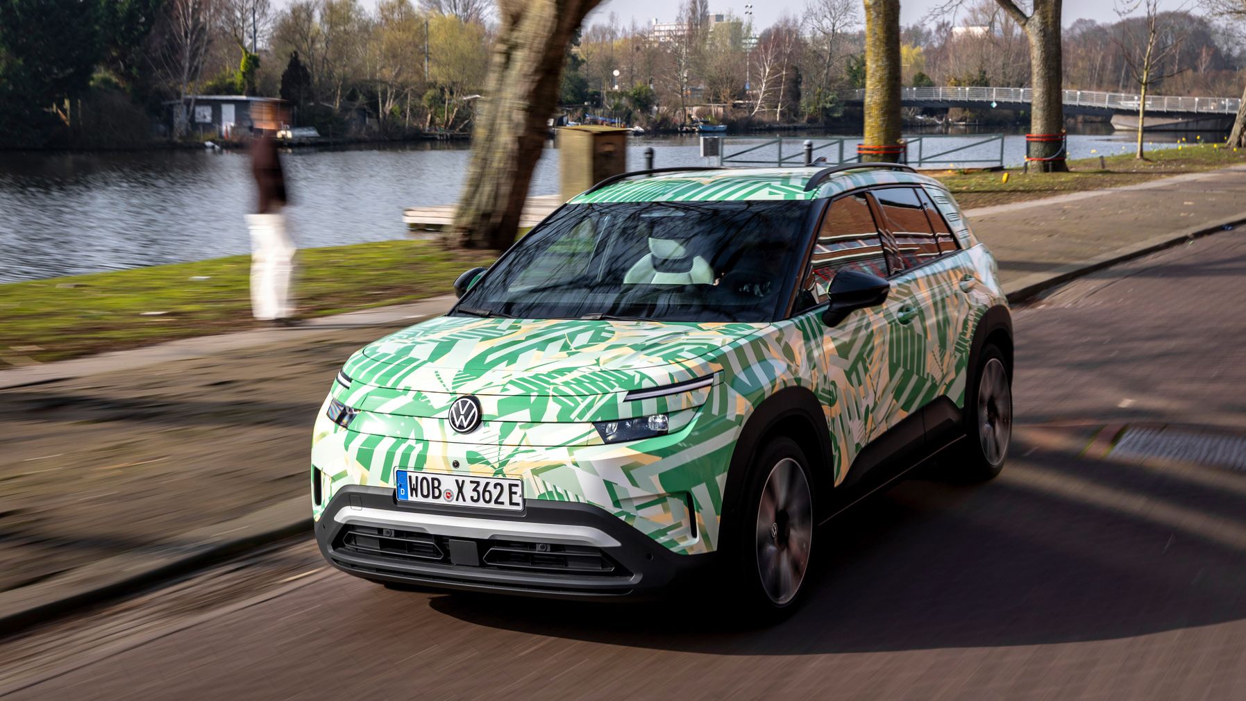 Ein Elektroauto fährt auf einer Straße entlang eines Flusses. Es hat eine grüne Tarnlackierung und moderne LED-Scheinwerfer. Im Hintergrund sind Bäume und eine Brücke zu sehen.