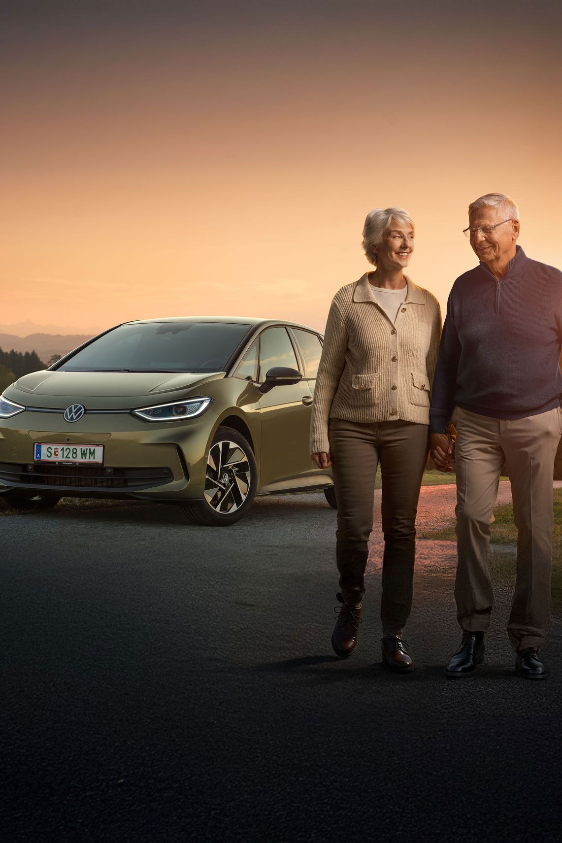 Ein älteres Paar spaziert zusammen auf einer Landstraße bei Sonnenuntergang. Im Hintergrund steht ein Auto, ein Volkswagen ID.3. Die Landschaft ist grün und hügelig.