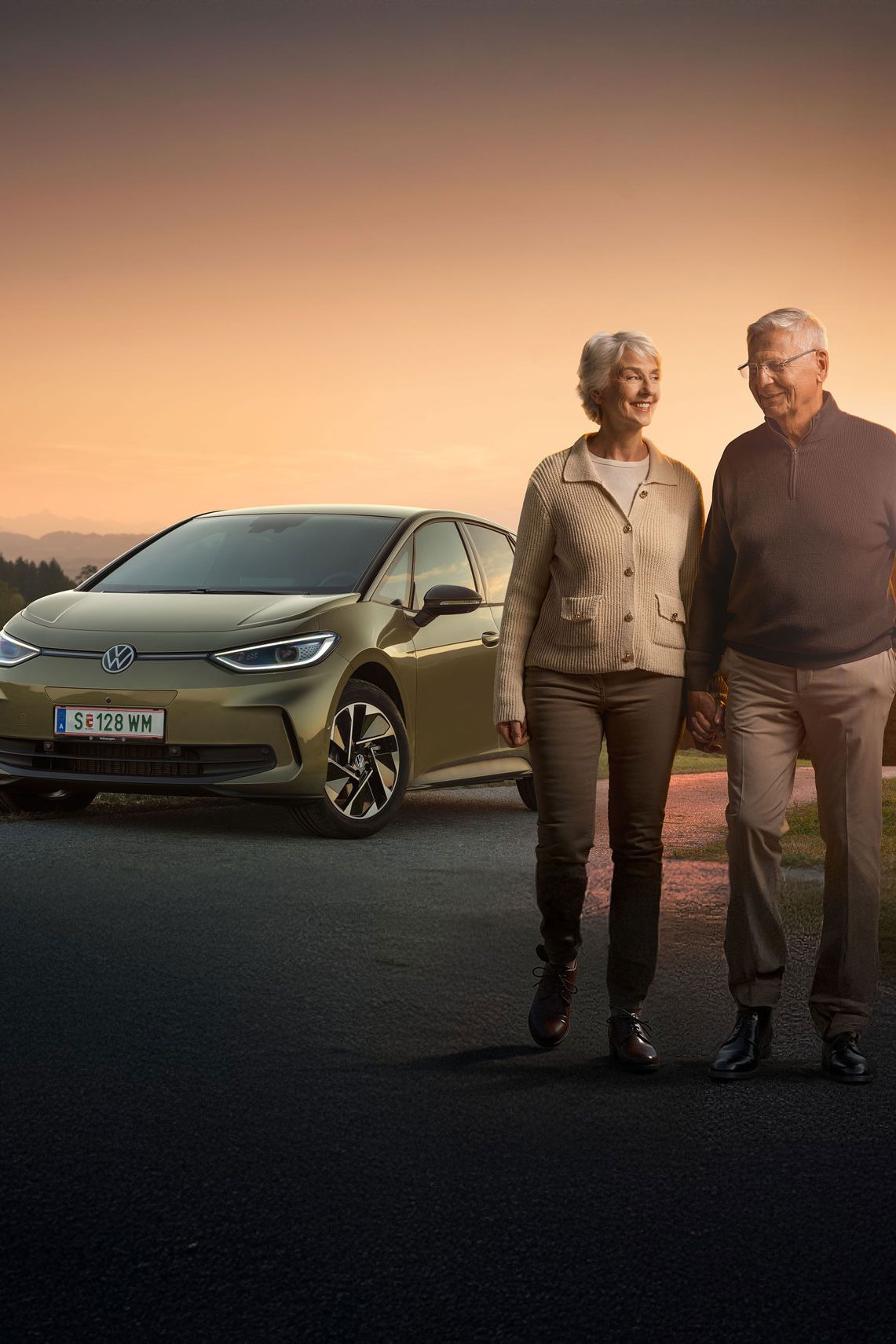 Ein älteres Paar spaziert zusammen auf einer Landstraße bei Sonnenuntergang. Im Hintergrund steht ein Auto, ein Volkswagen ID.3. Die Landschaft ist grün und hügelig.