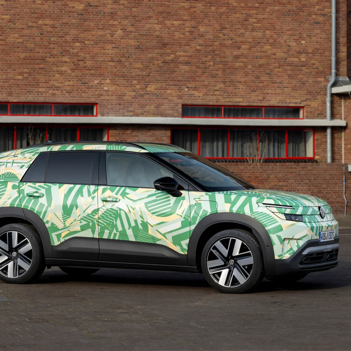 Ein SUV steht vor einem Backsteingebäude. Das Auto hat eine auffällige grüne und weiße Tarnlackierung. Die Fenster des Gebäudes sind rot umrahmt.