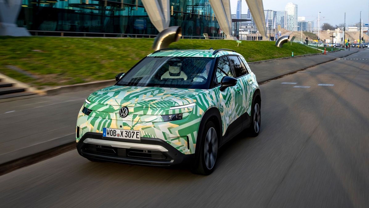 Ein Elektroauto fährt auf einer Straße neben einem modernen Gebäude mit schrägen Stützen und großer Glasfassade. Das Auto hat eine grüne und weiße Tarnlackierung. Im Hintergrund sind weitere Gebäude und eine graue, bewölkte Skyline zu sehen.