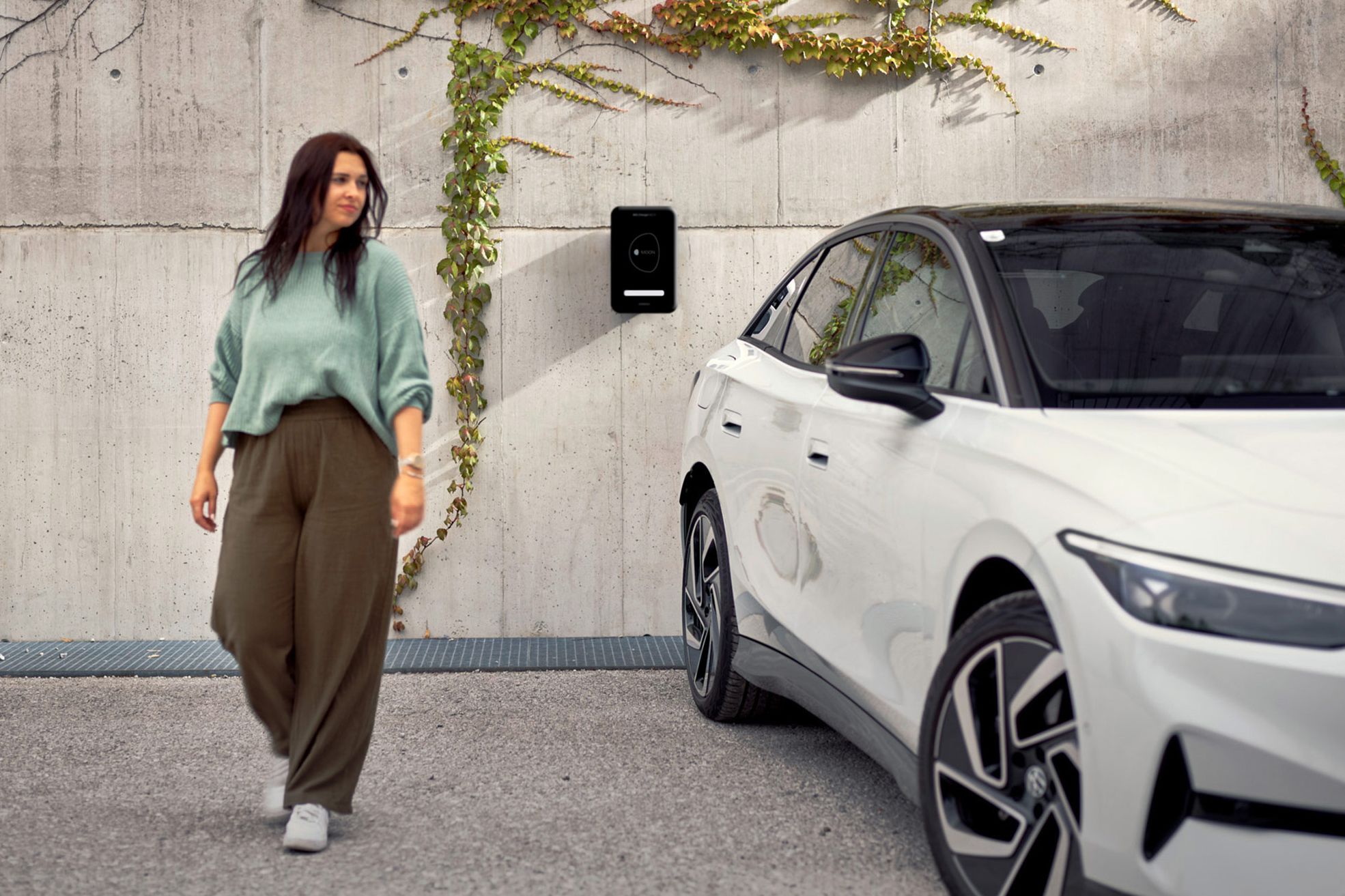 Eine Frau geht in der Nähe eines weißen ID.7, das an einer Wand mit einer Ladestation und Kletterpflanzen geparkt ist. Die Umgebung wirkt urban und modern.