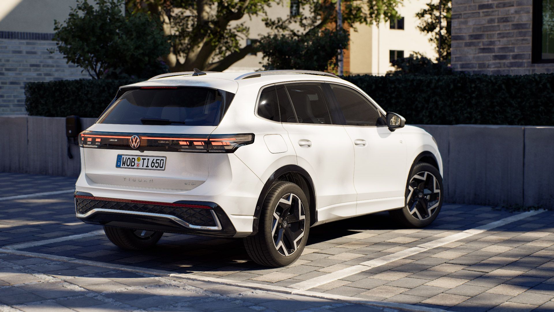 Weißer Volkswagen Tiguan steht schräg rückseitig in einem gepflasterten Parkplatz vor modernen Gebäuden und Büschen. Das Auto hat markante LED-Rückleuchten über die gesamte Heckbreite und schwarze Akzente an Dach, Felgen und Stoßfängern. Die Szene wirkt wie eine Werbeaufnahme bei Tageslicht.